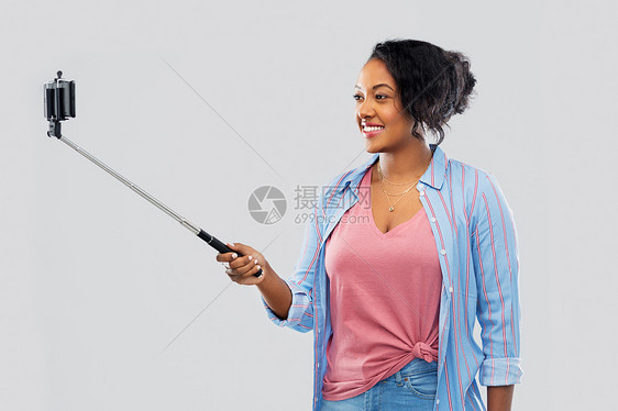 技术人的快乐的非裔美国妇女用智能手机灰色背景的自拍杆上拍照非裔美国妇女用智能手机自拍图片