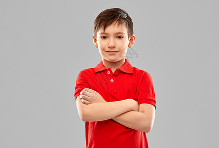 童,表情人的微笑的小男孩穿着红色马球T恤,交叉手臂灰色背景上微笑的男孩穿着红色的T恤,双臂交叉图片