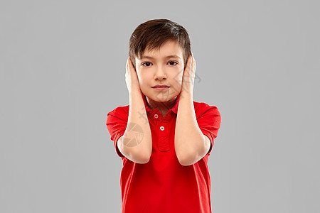 噪音,沉默人们的红色马球T恤小男孩的肖像,用手灰色背景上耳朵穿红色T恤的男孩用手闭着耳朵图片