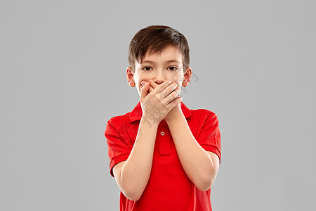 童,表情人的震惊的小男孩穿着红色马球T恤,用手灰色的背景上闭上嘴震惊的男孩穿着红色T恤,双手合嘴图片