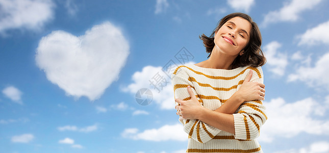 情人节,爱的人快乐的轻女人穿着条纹套头衫,闭着眼睛拥抱自己蓝天云的形状的心脏背景女人心形的云上拥抱自己图片