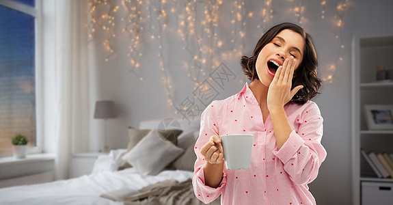 人们就寝时间的快乐困倦的轻妇女睡衣与咖啡打哈欠夜间卧室花环灯光背景快乐的哈欠轻的女人睡衣咖啡图片