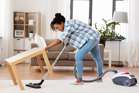 家庭家务快乐的非裔美国妇女家庭主妇与吸尘器清洁地毯下的桌子家里家里吸尘器的女人家庭主妇图片