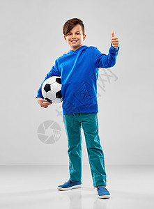 童,足球体育微笑的男孩穿着蓝色帽衫,足球拇指灰色背景带着足球的微笑的男孩竖大拇指图片