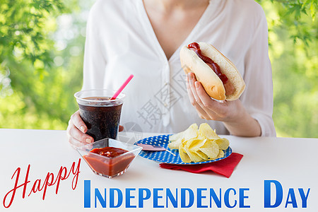 美国独立日,庆祝假日妇女抱着热狗可乐塑料杯与薯片番茄酱7月4日绿色自然背景特写女人用可口可乐吃热狗图片