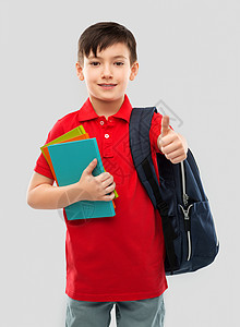学校,教育人的微笑的小学生男孩穿着红色马球T恤,戴着眼镜,带着书袋子,灰色背景上竖大拇指带着书书包的男孩竖图片