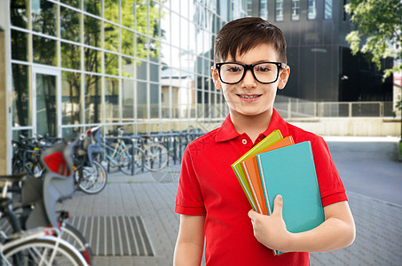 教育人的微笑的小学生男孩穿着红色马球T恤,戴着眼镜,学校院子背景上书带着书戴眼镜的微笑的小学生图片