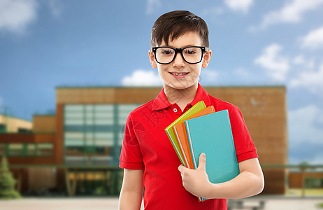 教育人的微笑的小学生男孩穿着红色马球T恤,戴着眼镜,带着学校背景的书带着书戴眼镜的微笑的小学生背景图片