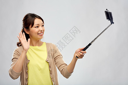 技术人的快乐的亚洲女人用智能手机自拍杆上拍照,并灰色背景上挥手亚洲女人用智能手机自拍图片