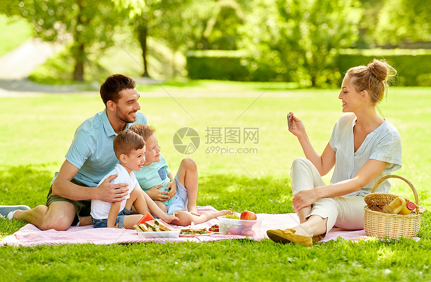 家庭休闲人的母亲夏季公园野餐时用智能手机拍摄快乐的父亲两个小儿子的照片妈妈公园野餐时给家人拍照图片