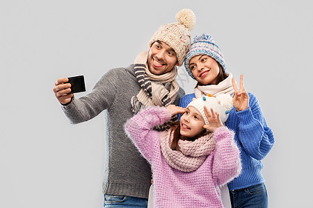 家庭,诞节技术快乐的母亲,父亲小女儿穿着冬天的衣服,用智能手机灰色背景下自拍幸福的家庭用智能手机自拍图片