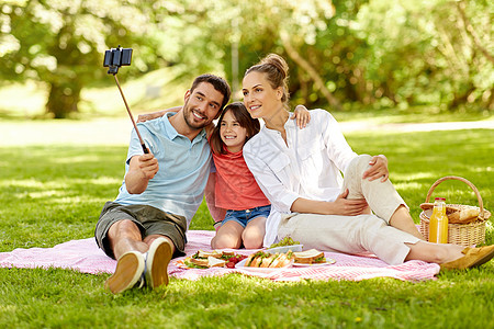 家庭休闲技术快乐的母亲父亲女儿夏季公园用智能手机自拍杆上野餐拍照家人公园野餐自拍图片