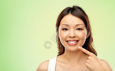 美丽人的快乐微笑的轻亚洲妇女指着她的嘴牙齿石灰绿色的自然背景快乐微笑的亚洲轻女人抚摸她的脸图片
