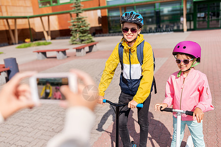 教育,童人的快乐的学校孩子戴着头盔,背包滑板车被智能手机拍摄户外学校的孩子们用滑板车牢房里拍照图片