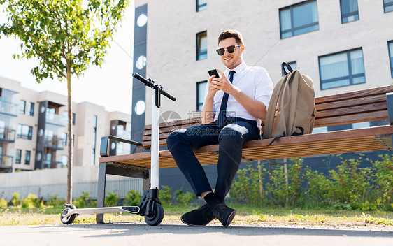 商业人轻的微笑商人与智能手机,背包滑板车坐城市的街道长凳上拥智能手机背包滑板车的商人图片