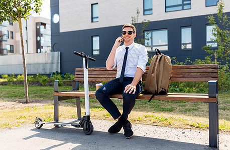 商业人轻的微笑商人,背包滑板车坐街上的长凳上,给城市的智能手机滑板车的商人智能手机上打电话图片