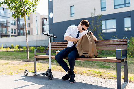 商业人轻的商人,带着袋子电动滑板车,咖啡外卖纸杯里,坐城市的长凳上城里包滑板车咖啡的商人图片