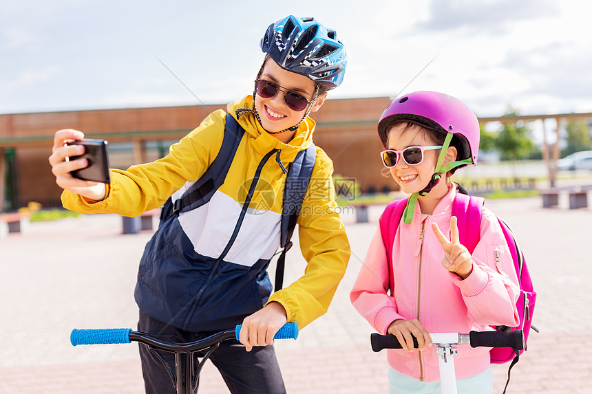 教育,童人的快乐的学校孩子戴着头盔,背包骑着滑板车,户外用智能手机自拍快乐的学校孩子与滑板车自拍图片