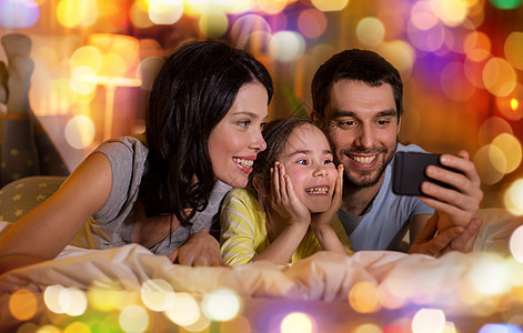 人们家庭的快乐的母亲,父亲小女儿,晚上家里睡觉时带着智能手机快乐的家庭,晚上床上智能手机图片