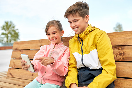 童技术人的快乐的孩子兄弟姐妹智能手机坐户外的木街长凳上孩子们着智能手机坐长凳上图片