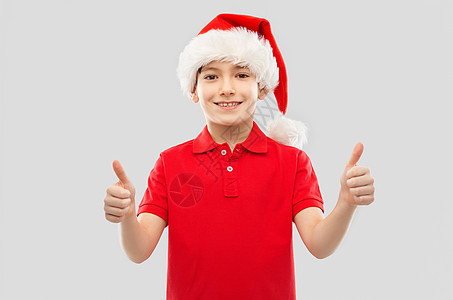 诞节,假日童的微笑的小男孩穿着红色马球T恤诞老人助手帽子,灰色背景上竖大拇指戴着诞老人帽子的微笑男孩竖图片