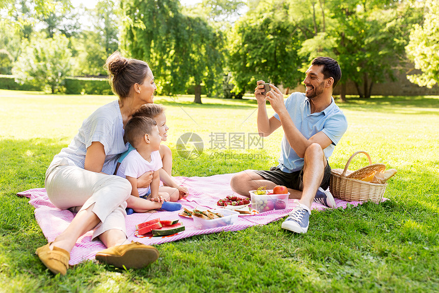 家庭休闲人的父亲夏季公园野餐时用智能手机拍下了幸福的母亲两个小儿子的照片父亲公园野餐时给家人拍照图片