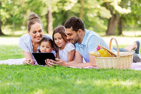 家庭休闲人的快乐的母亲,父亲两个女儿,用平板电脑铺设夏季公园的野餐毯上家庭与平板电脑夏季公园野餐图片