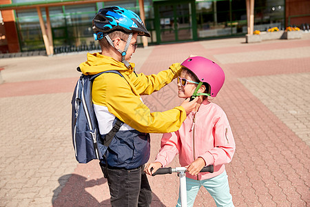 安全,童护理快乐的学校男孩与背包紧固女孩的头盔滑板车骑学校男孩为滑板车系紧女孩的头盔图片