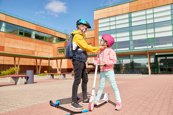 安全,童护理快乐的学校男孩与背包紧固女孩的头盔滑板车骑学校男孩为滑板车系紧女孩的头盔图片