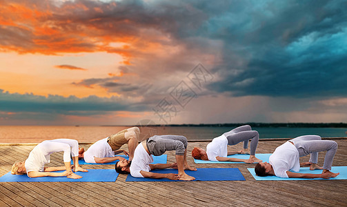 健身,运动,瑜伽健康的生活方式群人日落背景下的海上码头上桥式群人户外瑜伽练图片