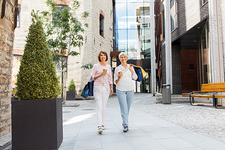 销售,消费主义人的两个老年女子朋友带着购物袋,喝着外卖咖啡,沿着塔林市街走城里购物袋咖啡的高级女人图片