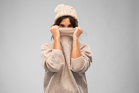 冬天的季节人们的快乐的轻女人戴着针帽子,脸上覆盖着灰色背景上温暖的毛衣穿着针冬季毛衣帽子的轻女人图片