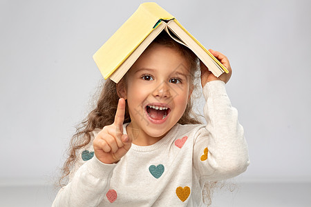 阅读,教育童的微笑的小女孩的肖像,头上书,屋顶上灰色的背景带着书的微笑女孩的肖像图片
