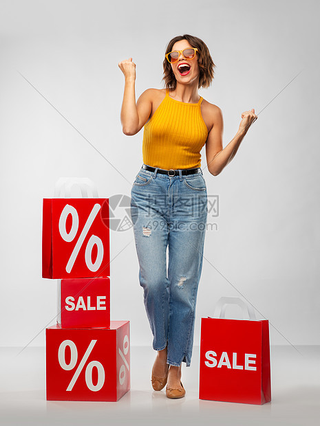 销售,消费主义人们的快乐的微笑轻妇女戴太阳镜,芥末黄色上衣牛仔裤与购物袋庆祝成功的灰色背景带着购物袋的快乐微笑的图片