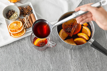 诞节季节饮料的手与瓢浇热覆盖葡萄酒与橙色片芳香香料璃杯灰色背景用勺子把热覆盖的葡萄酒倒入璃杯图片