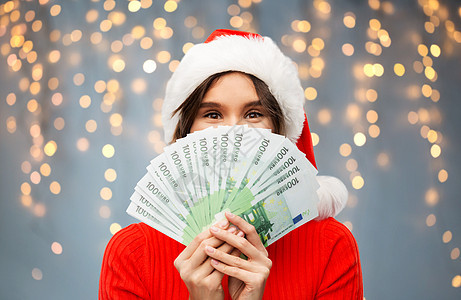 诞节,假日金融快乐的轻女人戴着诞老人的帽子,躲灰色背景上的节日灯上的欧元钞票后诞快乐的女人戴着诞帽带着图片