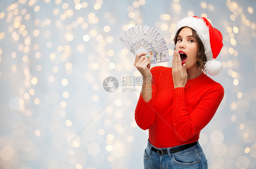 ‘~诞节,假日金融惊讶的轻妇女戴着诞老人的帽子,着美元的钞票节日的灯光背景带着钱穿着诞老人帽子的惊讶表妹  ~’ 的图片