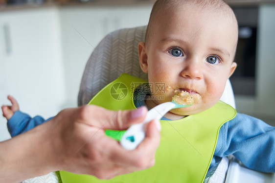 微笑的男孩家里的高椅子上被母亲用勺子喂固体食物的肖像图片