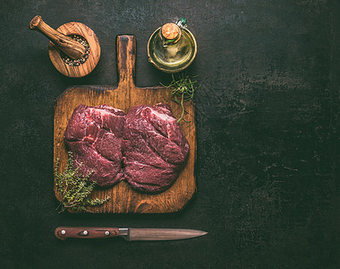 原始大理石牛肉木制切割板上,香草,香料,油刀黑暗的乡村背景,顶部视图为您的烹饪食谱图片