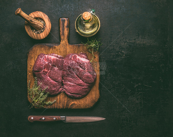 原始大理石牛肉木制切割板上,香草,香料,油刀黑暗的乡村背景,顶部视图为您的烹饪食谱图片