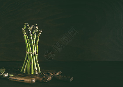 绿色芦笋站黑暗的乡村餐桌背景与木制切割板刀图片