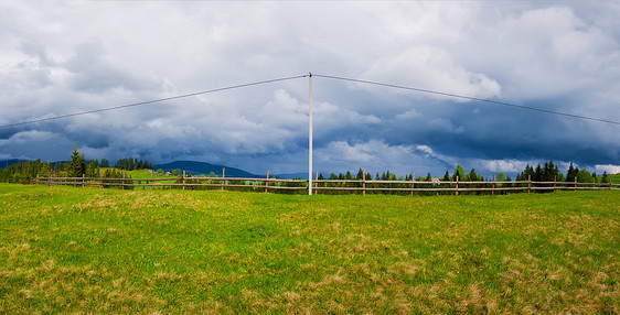 电力传输杆山上,电缆沿木轨围栏通向村庄全景景观,绿色的田野多云的天空背景图片