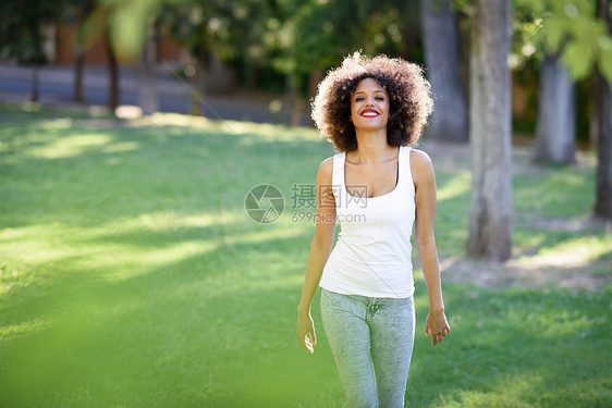 轻的黑人妇女,留着AFRO发型,城市公园微笑混合女孩穿着白色T恤蓝色牛仔裤草地上散步图片