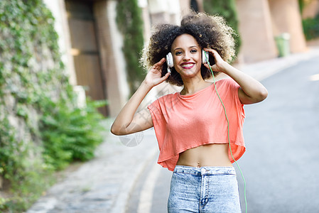 城市背景中轻迷人的黑人女孩的肖像,用耳机听音乐女人穿着橙色T恤蓝色牛仔裤,留着AFRO的发型图片
