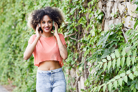 城市背景中轻迷人的黑人女孩的肖像,用耳机听音乐女人穿着橙色T恤蓝色牛仔裤,留着AFRO的发型图片