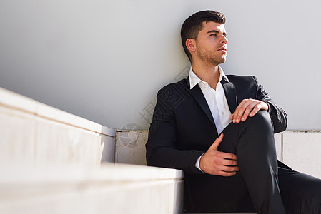 轻的商人现代办公楼附近,穿着黑色西装白色衬衫坐地板上城市背景中蓝眼睛的男人图片