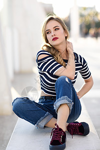碳纤金发女人,时尚模特,坐城市背景的长凳上漂亮的轻女孩穿着条纹T恤蓝色牛仔裤街上漂亮的俄罗斯女,辫子背景