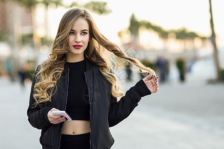 金发女人城市背景下用她的智能手机发短信漂亮的轻女孩穿着黑色夹克街上散步漂亮的俄罗斯女,留着长长的波浪发型背景图片