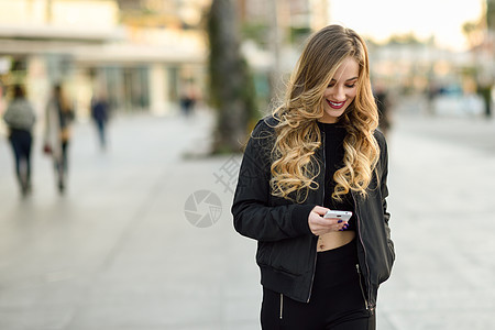 金发女人城市背景下用她的智能手机发短信漂亮的轻女孩穿着黑色夹克街上散步漂亮的俄罗斯女,留着长长的波浪发型背景图片