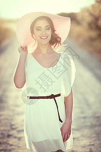 春天的条乡村道路上闭着眼睛的轻女人的肖像穿着白色连衣裙,棕色腰带粉红色太阳帽的女孩背光摄影与太阳女人的后图片